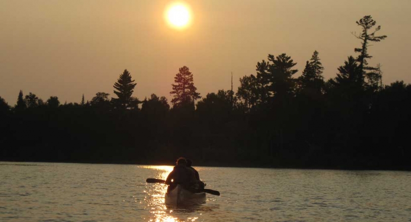 the silhouette of a canoe appears on calm water, with the sun just above the tree-line in the background.